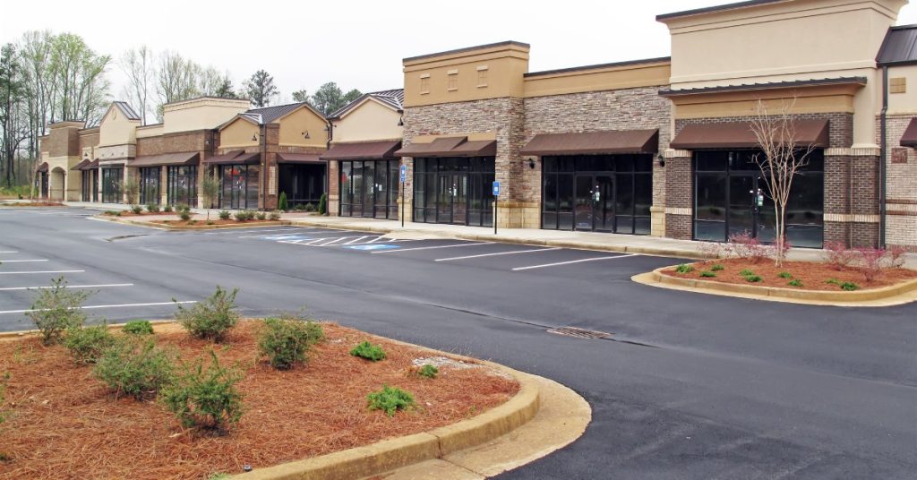 A strip mall parking area with clearly defined parking spots. There are median areas filled with red mulch and small shrubs.