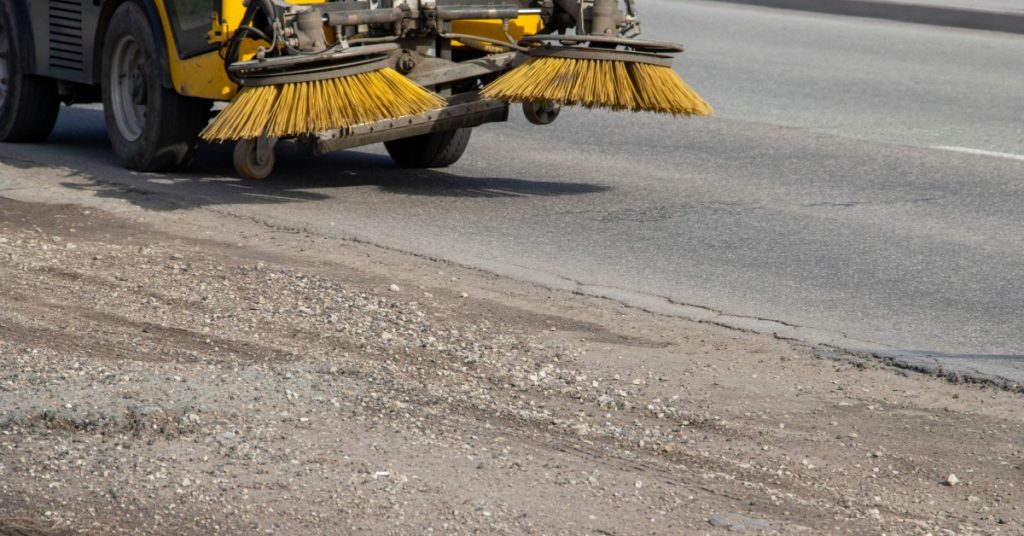 An asphalt construction site with a large industrial sweeper driving across it. It has large yellow sweepers.