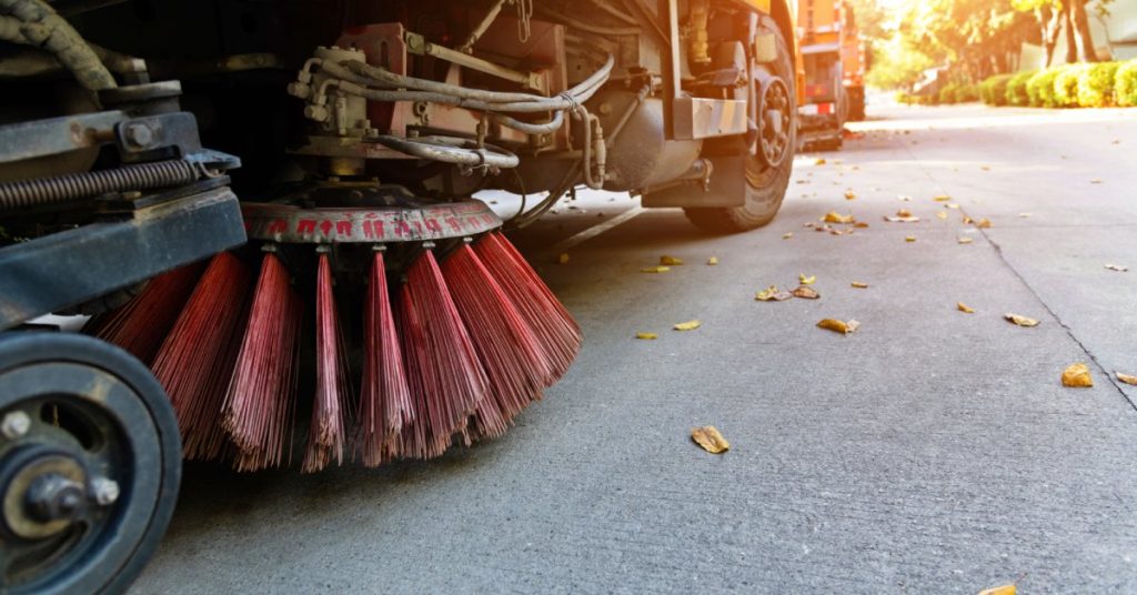 Multiple street sweeper trucks are going over pavement. The focus is on a large, red sweeper brush.