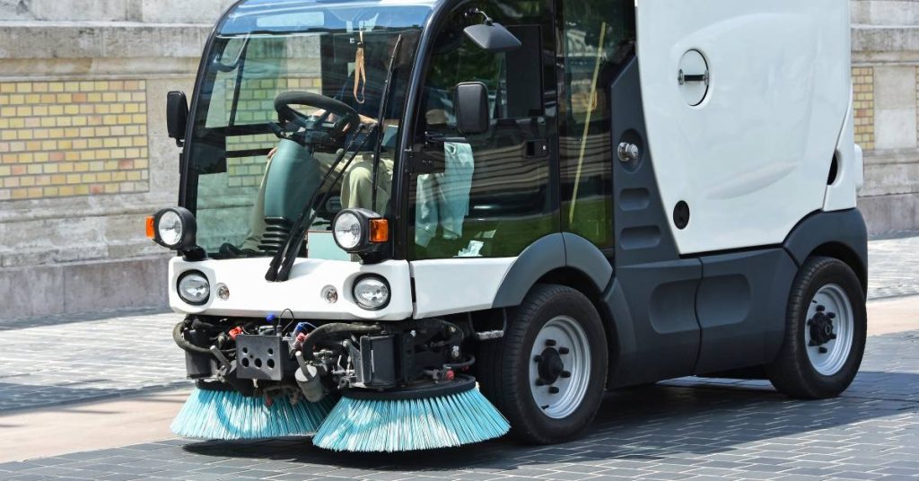 A small white street sweeper with green brushes in the front is driving through a narrow, gray brick road.