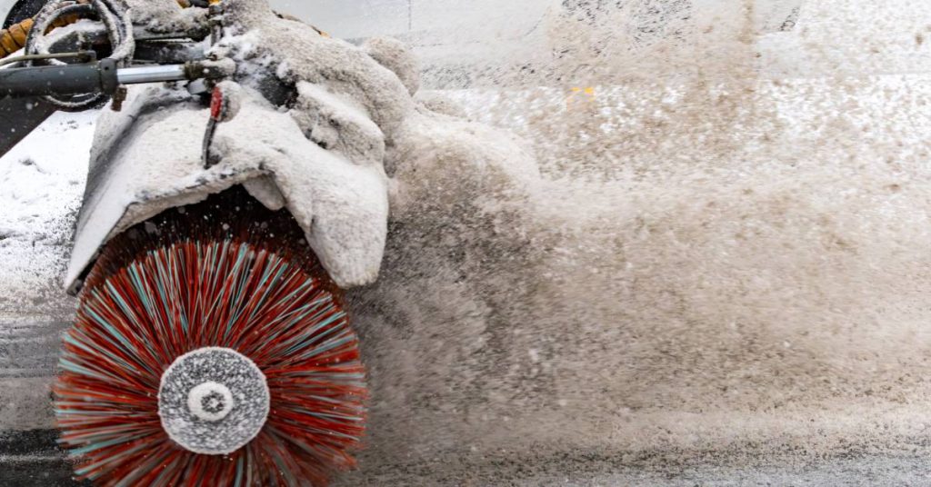 A spinning street sweeper brush in the middle of a snowstorm. The sweeper is blowing snow off to the side.