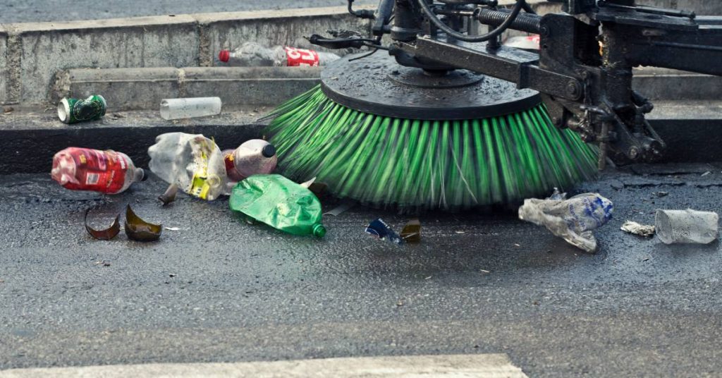  A large green spinning brush of a street sweeper. It's passing through garbage containing several plastic bottles.