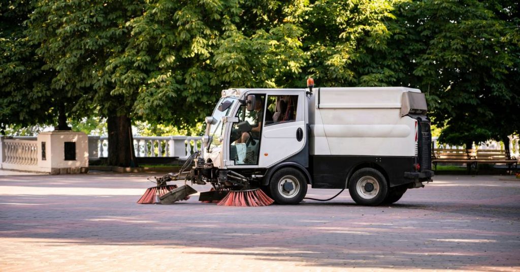 A small white street sweeper with large red brushes is sweeping through the road in a residential area.