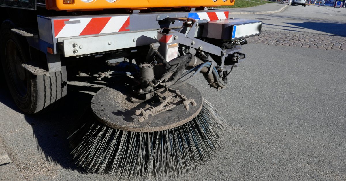 A large orange street sweeper with a front-facing circular brush drives through a public road.