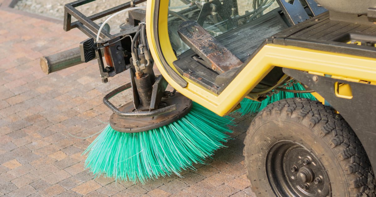 A small yellow sweeper vehicle with green circular brushes sweeps through a paved surface in a construction site.