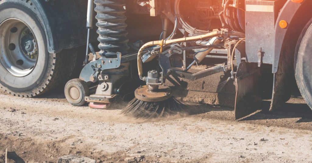 A road sweeper with a small circular brush going through a dirty construction site. The brush is sweeping up dust.