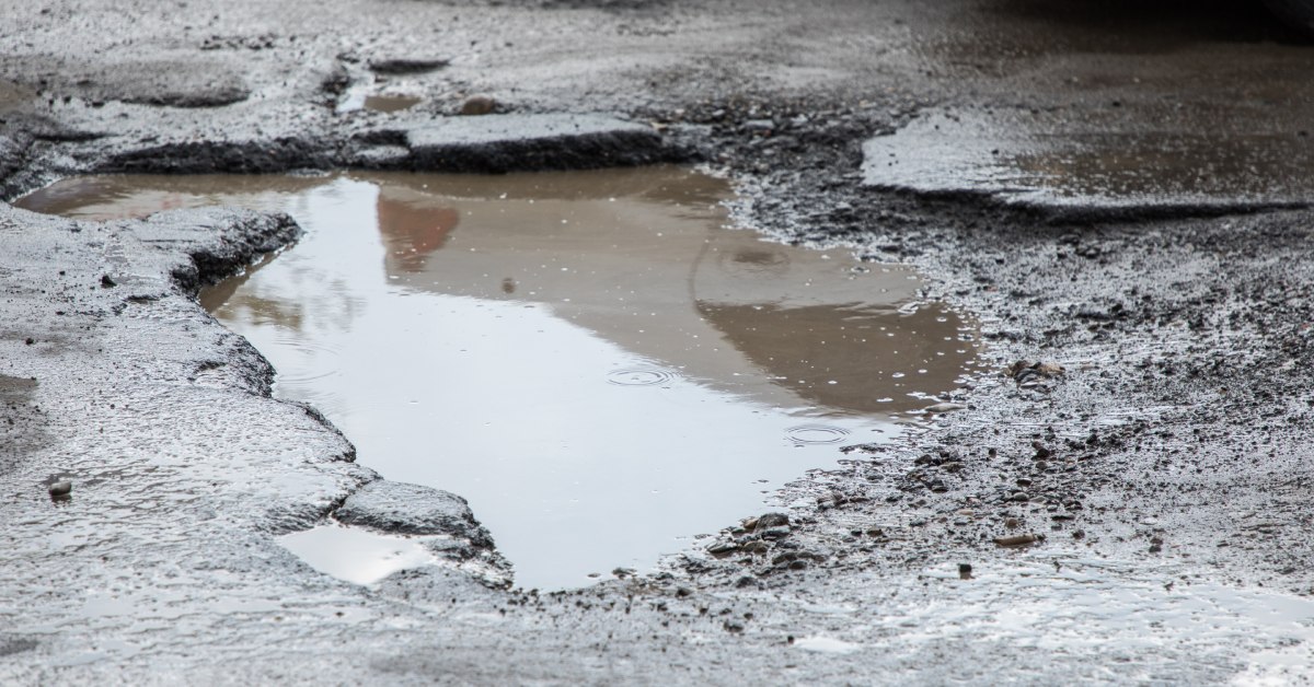 Closeup of a wet patch of asphalt with a large pothole in it. The hole is full of dirty brown water.