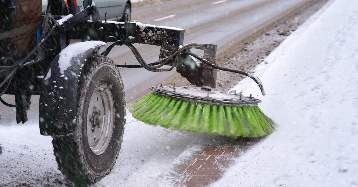 A sweeper with a large green spinning brush attachment is driving down a road and clearing snow off of the side.