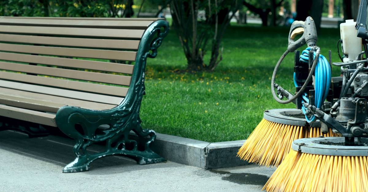 A grassy area surrounded by a concrete barrier. There is a bench to the left and the brushes from a street sweeper on the right.