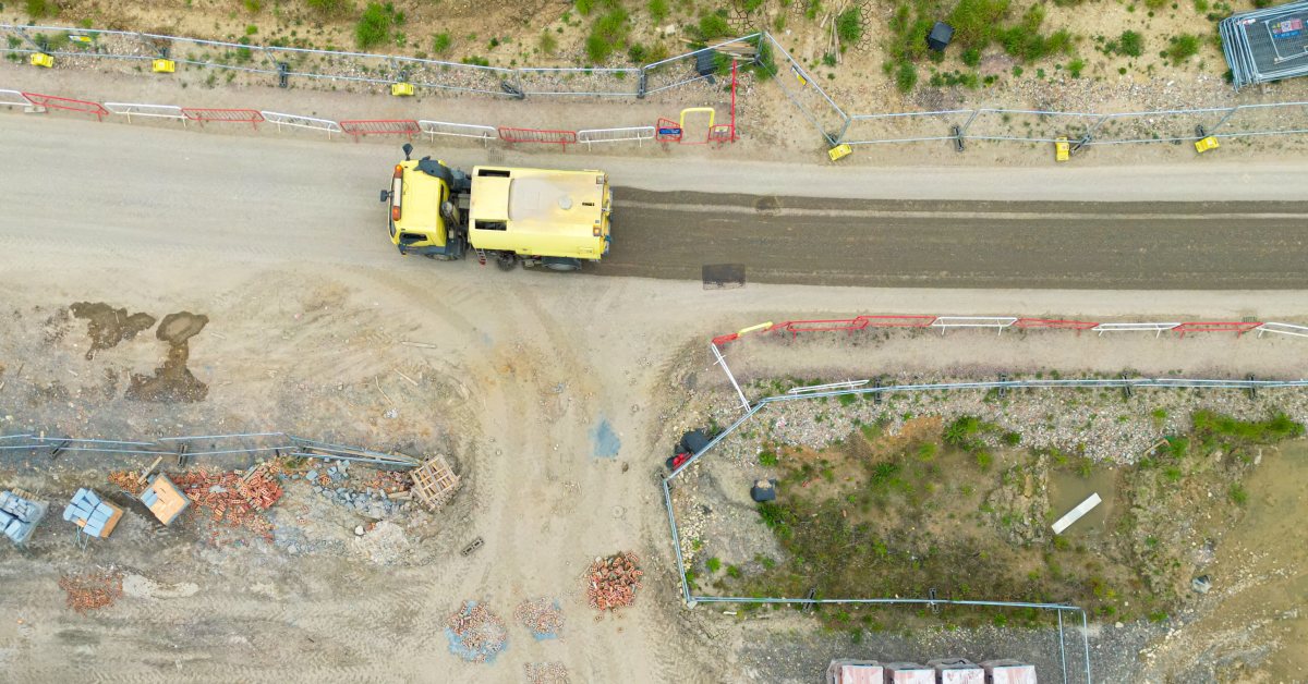 Top down aerial view of a construction site. A yellow vehicle is driving along a dirty road, leaving a clean streak behind it.