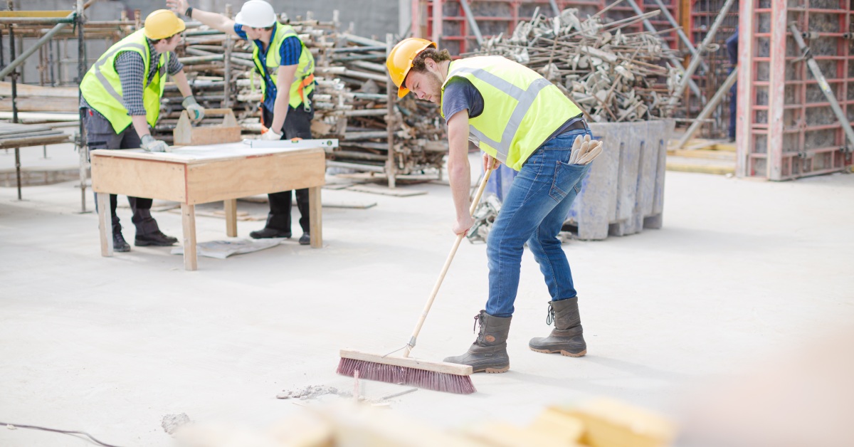 Man at a construction site is wearing a bright vest and hard hat while sweeping the floor with a large push bristle broom.