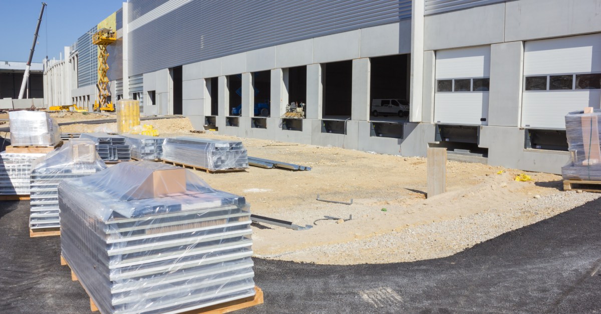 A construction site outside of a warehouse in progress. Several nearly stacked pallets are wrapped up out in front of it.