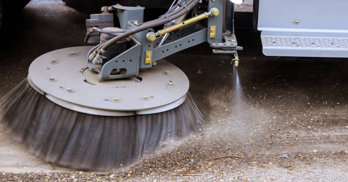 Industrial street sweeper with a large gray brush. The sweeper is spraying water down onto the asphalt ahead of the brush.