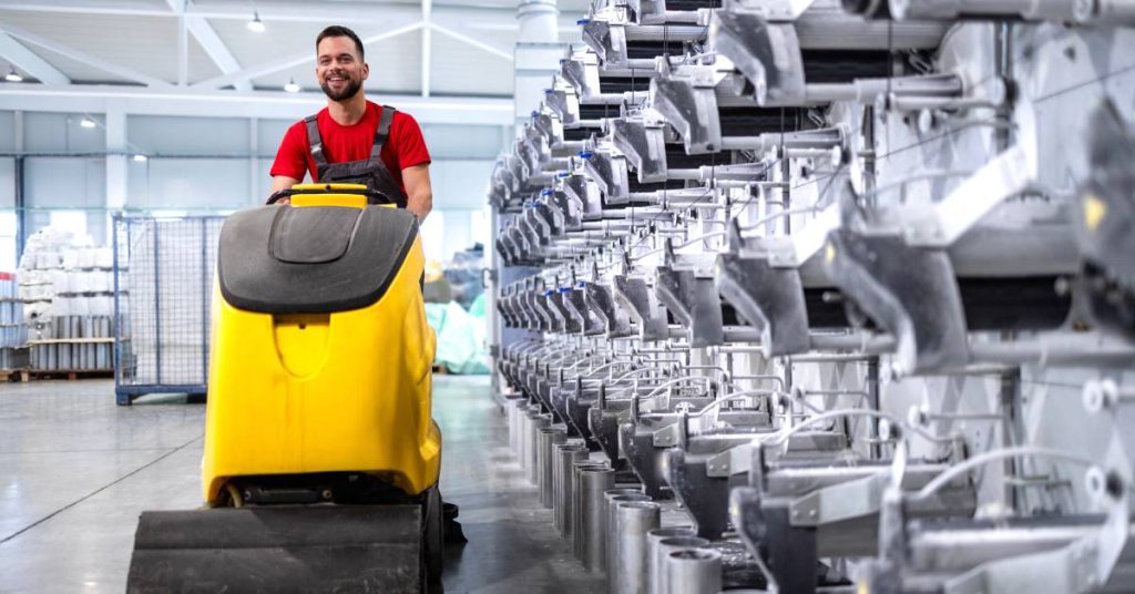 A man in a red shirt riding a yellow floor sweeper through a warehouse. Large racks of auto parts are visible on the side.