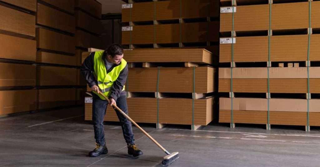 Young man wearing a bright yellow work vest sweeping a dirty floor. Behind him are several stacked wooden pallets.