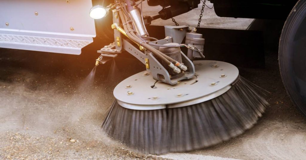  Street sweeper brushing along the edge of a road. The brush is kicking up a large cloud of dust and dirt.