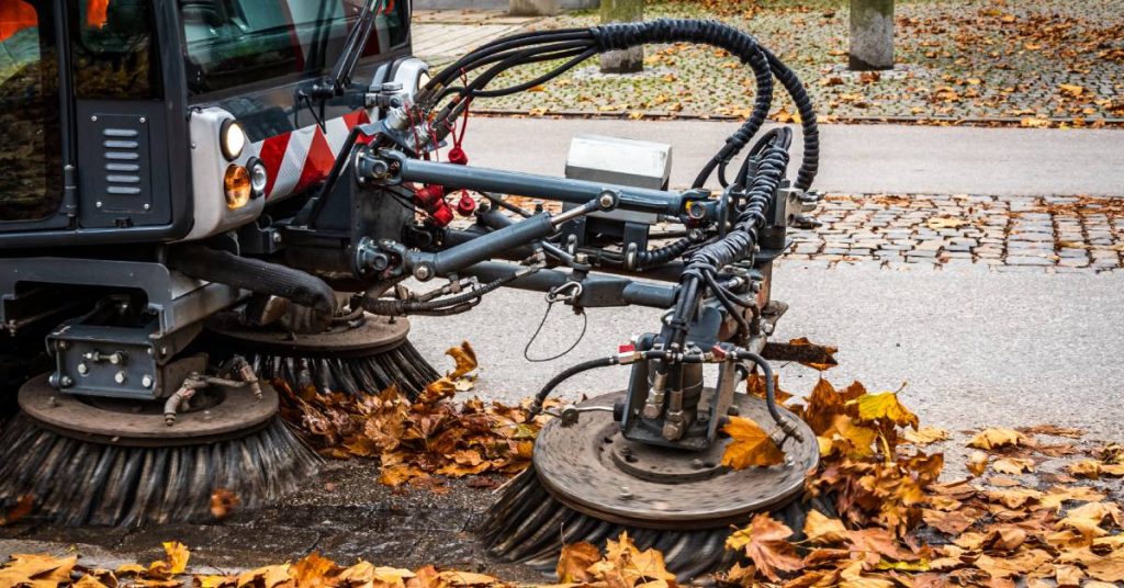 Street sweeper with extended brushes moving along a sidewalk. The brushes are moving several dead leaves out of the way.