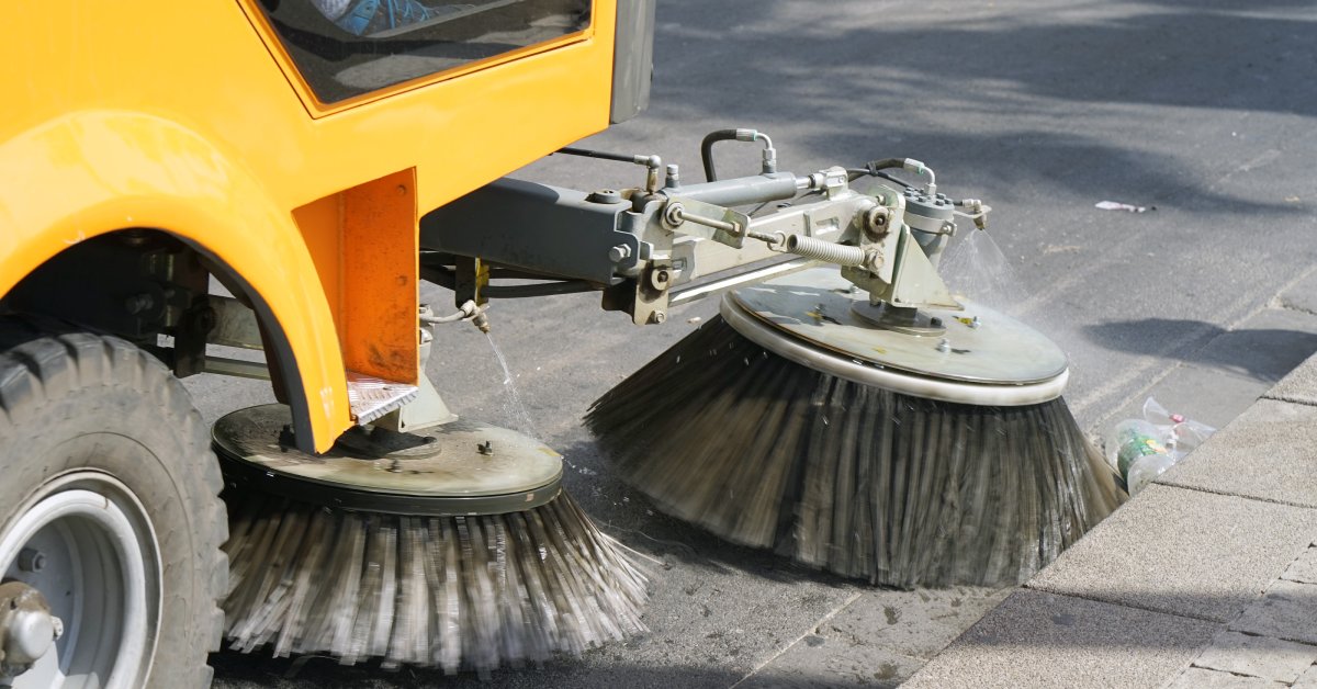 A street sweeping machine with a yellow chassis and round bristle brushes. It clears debris from asphalt pavement.