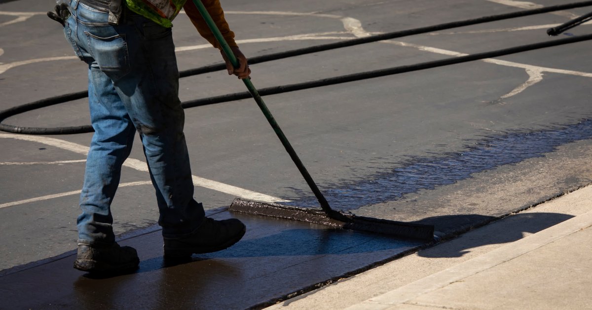 A man wearing jeans and boots applies a black sealcoat to asphalt pavement with a sealcoating brush.