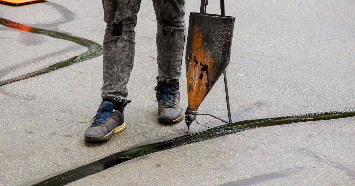 A man in black jeans and tennis shoes using metal equipment to fill pavement cracks with black sealant.