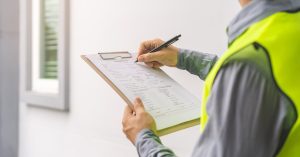 A man in a safety vest holds a clipboard with a paper form in one hand and a black pen in the other.