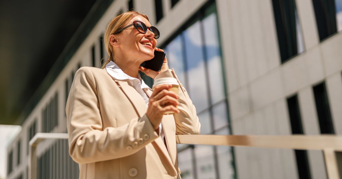 A woman wearing a tan suit jacket and sunglasses smiles and holds a coffee cup while talking on a cell phone.