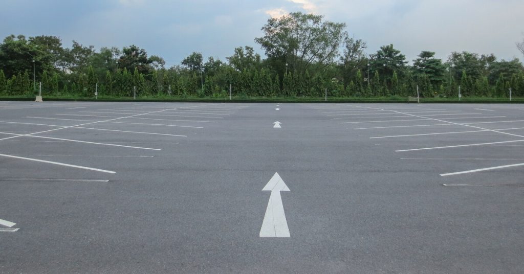 A gray parking lot with white arrows pointing forward and white lines for parking spaces sits empty.