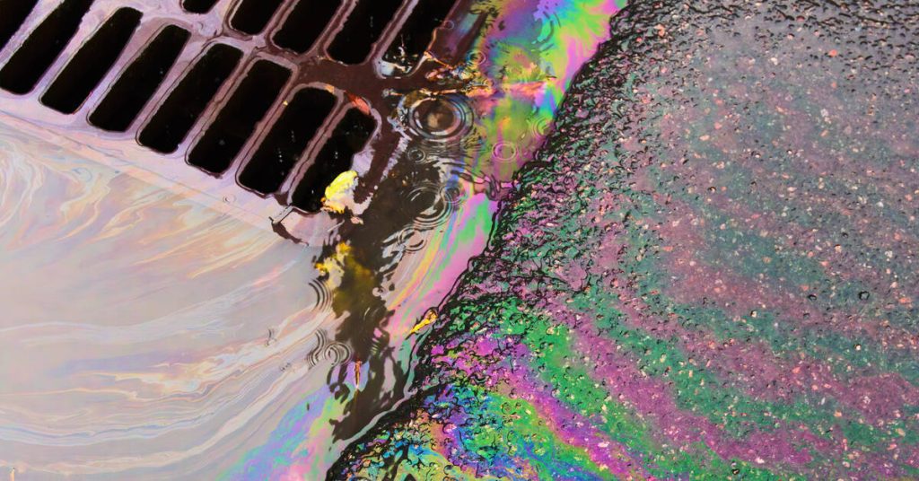 Oil mixed with water creates colorful ripples as it flows into a metal drain on the parking lot ground.