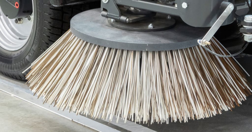 A round brush in front of a wheel on an industrial sweeping machine hovers above paved facility flooring.