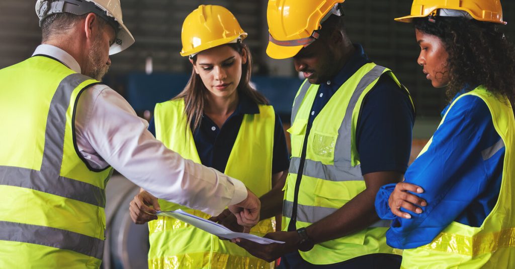 Four warehouse workers in yellow safety vests and hard hats look at a paper together. One of them points at the paper.