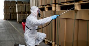 A person wearing a protective suit and a face covering sprays a solution amongst the boxes stacked in a warehouse.