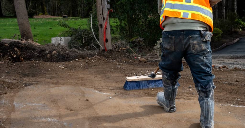 A construction worker in blue jeans and boots uses a broom with blue bristles to sweep the dirt floor.