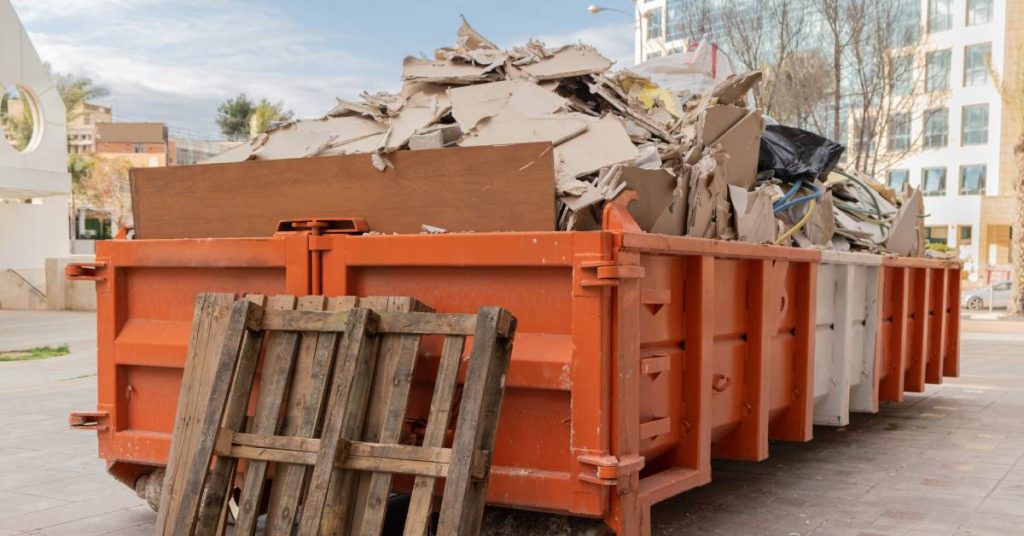 A large orange dumpster is full of pieces of wood and cardboard. A wood pallet leans against the dumpster.