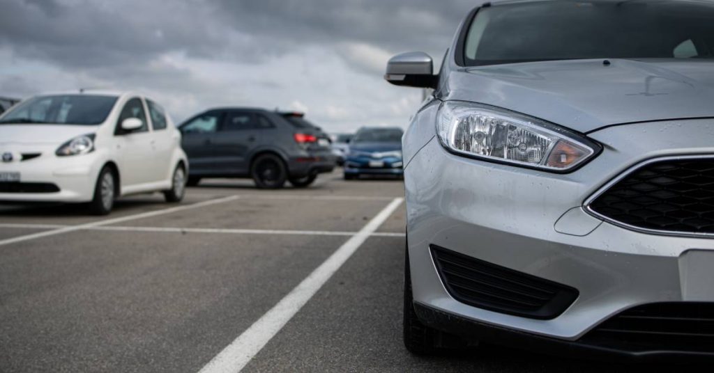 A silver car is parked in a parking space. There are several other parking spaces and cars behind it, and the sky is gray.