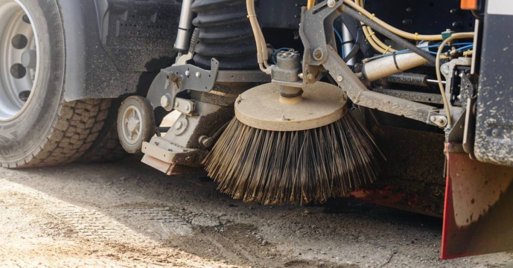 A round bristle brush near the wheels of a street sweeping machine hovers over a spot of broken asphalt pavement.