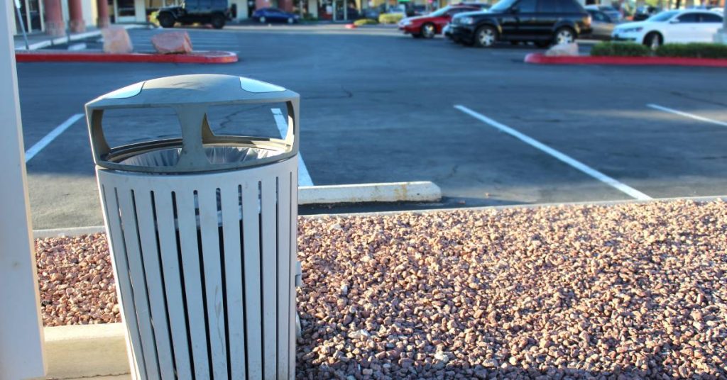 A white trash can with a cover sits surrounded by gravel next to a parking lot with cars in a shopping strip.