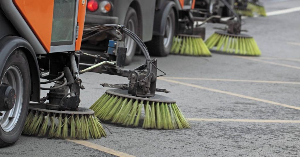 A row of orange street sweeping machines with yellow round bristle brushes sit parked on the pavement.