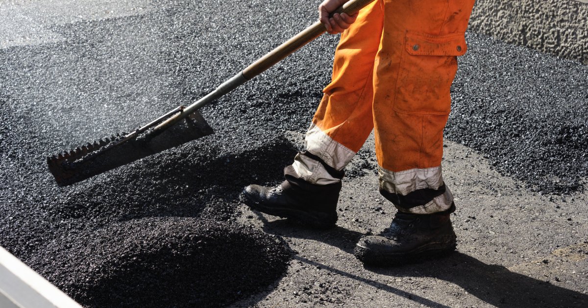 A worker in orange pants and black boots rakes over fresh black asphalt pavement outside during the day.