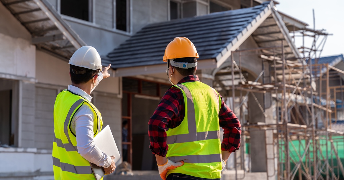 Two construction workers in yellow safety vests and hard hats stand looking at a house they are building.