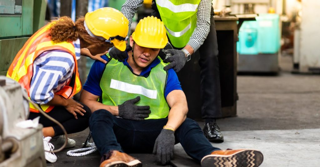 An industrial worker sits on the ground with his hand on his stomach while two other workers check on him.