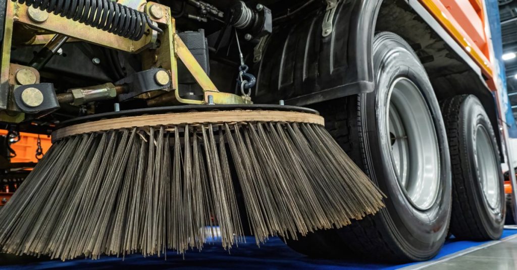The round black bristles of a large street sweeping machine hover above the ground inside an industrial facility.
