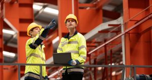 Two industrial workers in yellow safety jackets and hard hats stand with a laptop in a manufacturing facility.