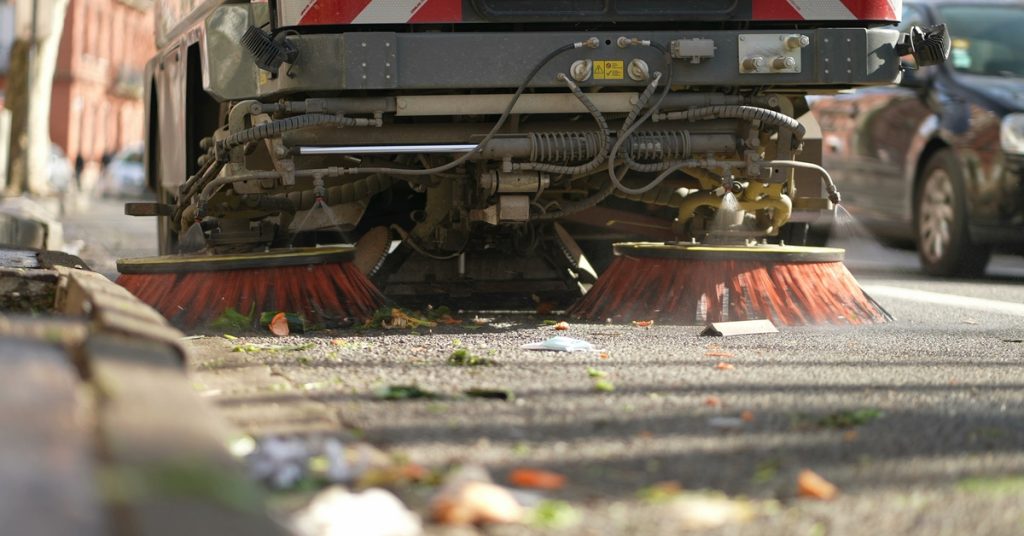 Large round red bristle brushes at the front of a street sweeper vehicle spin to clean the road with debris on it.
