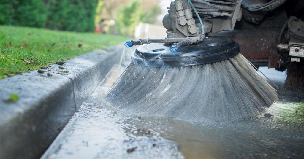 A stream of water comes out of a jet next to the round brush of a street sweeper truck to help in the cleaning process.