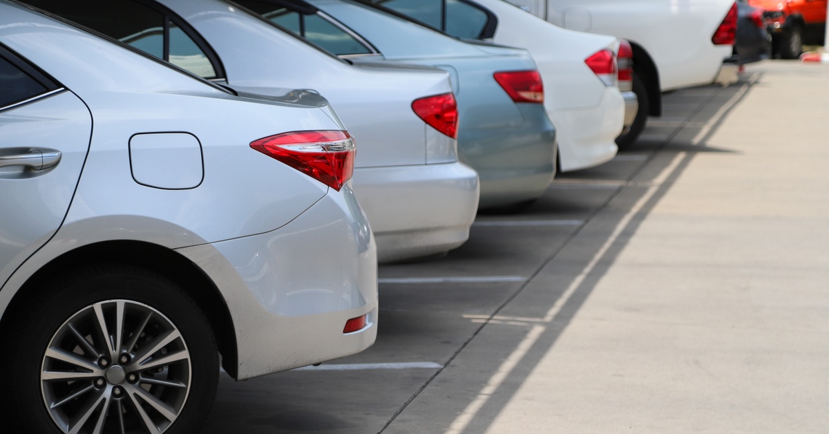 Several gray and white cars are lined up next to each other in a shaded section of a parking lot during the day.