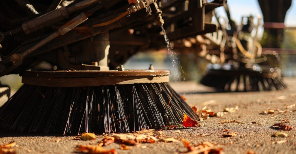 The round black bristle brushes of a street sweeper vehicle press against the ground to clean leaves off of pavement.