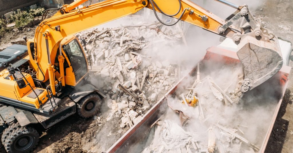 A large yellow excavator machine drops debris into a truck, sending dust flying through a construction site.