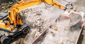 A large yellow excavator machine drops debris into a truck, sending dust flying through a construction site.