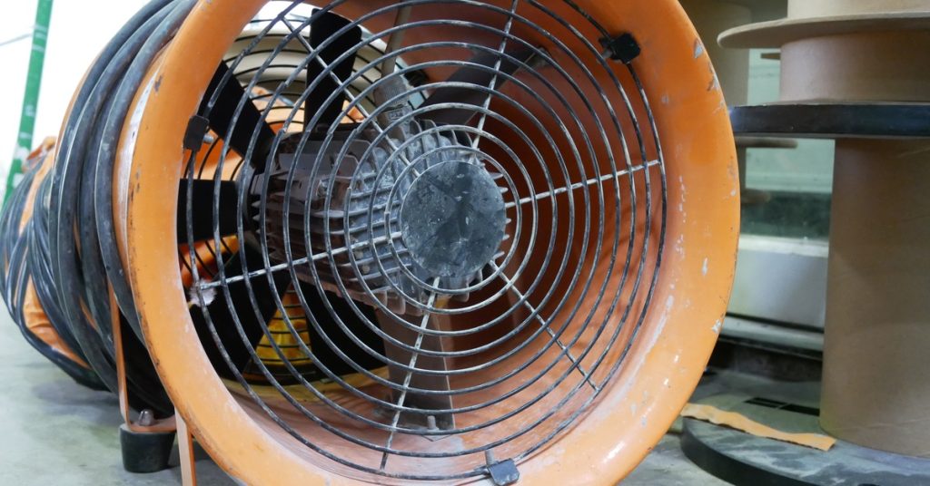 An orange portable exhaust fan with its cord wrapped up sits on the dusty concrete floor of a construction site.