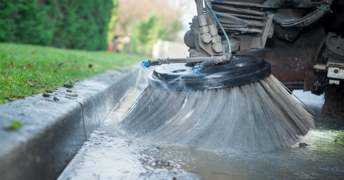 The gray circular bristle brush of a street sweeping truck cleans along the edge of a road while spraying water.
